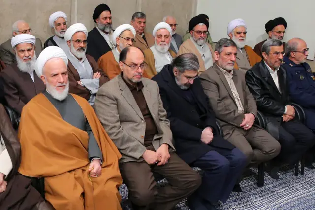 Mahmoud Ahmadinejad oturuyor. Üç sıra boyunca oturmuş yirmiye yakın erkek var.