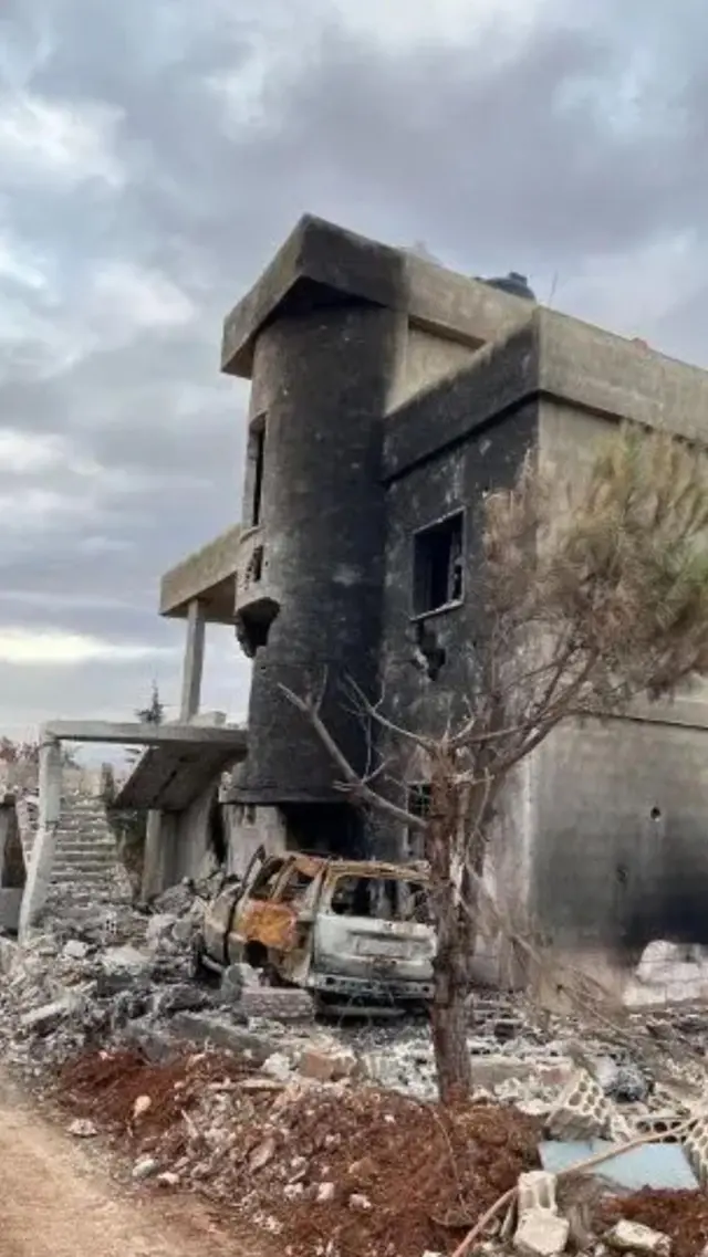 Bombalanmış bir ev. Duvarları is içinde. Önünde hurdaya dönmüş bir araba var.
