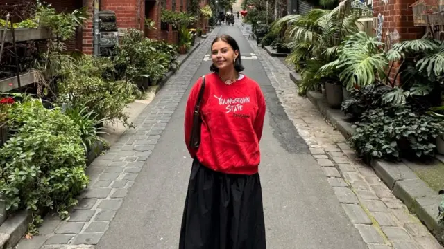 Uzun ince bir yolda tek başına duran genç bir kadın ellerini arkasına bağlamış. Üzerinde kırmızı bir kazak var.